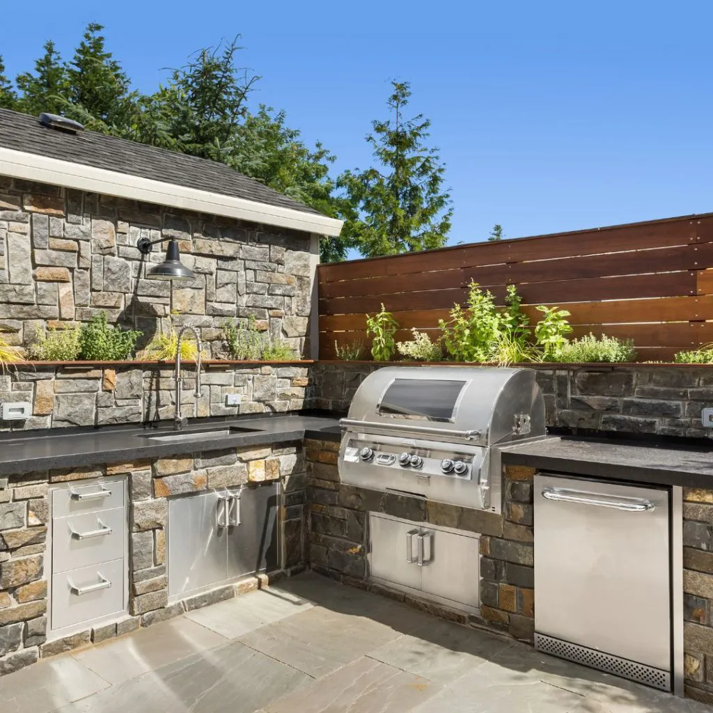 Outdoor stone kitchen with stainless steel appliances, including a grill and sink, surrounded by greenery, constructed by Mr. Pavers Contractor Services, Inc. in Rodeo.