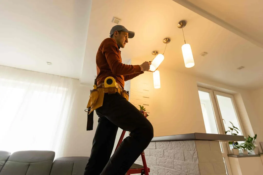 Electrician on a ladder installing pendant lights in a modern living space, wearing a tool belt.