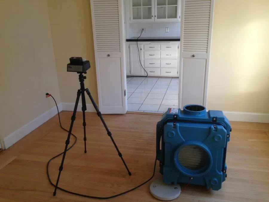 A blue air filtration device and a camera on a tripod are set up on a wooden floor, facing a kitchen area with white cabinets.