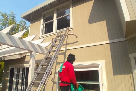 A window cleaner from NorCal Maintenance Inc. working on the exterior of a two-story house in Windsor, standing on a ladder with a bucket, showcasing professional cleaning services.