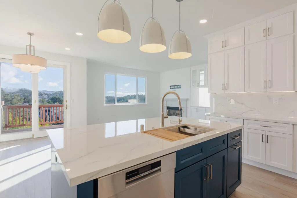 A modern kitchen with a marble island, navy blue cabinets, and white cabinetry, featuring pendant lights and a large window view in San Jose, designed by NuHome Remodeling.