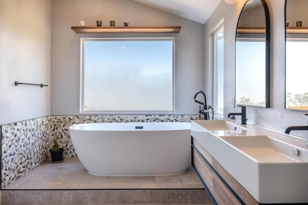 A modern bathroom featuring a freestanding bathtub, dual sinks with sleek faucets, a large frosted window, and decorative tiles on the walls and floor.