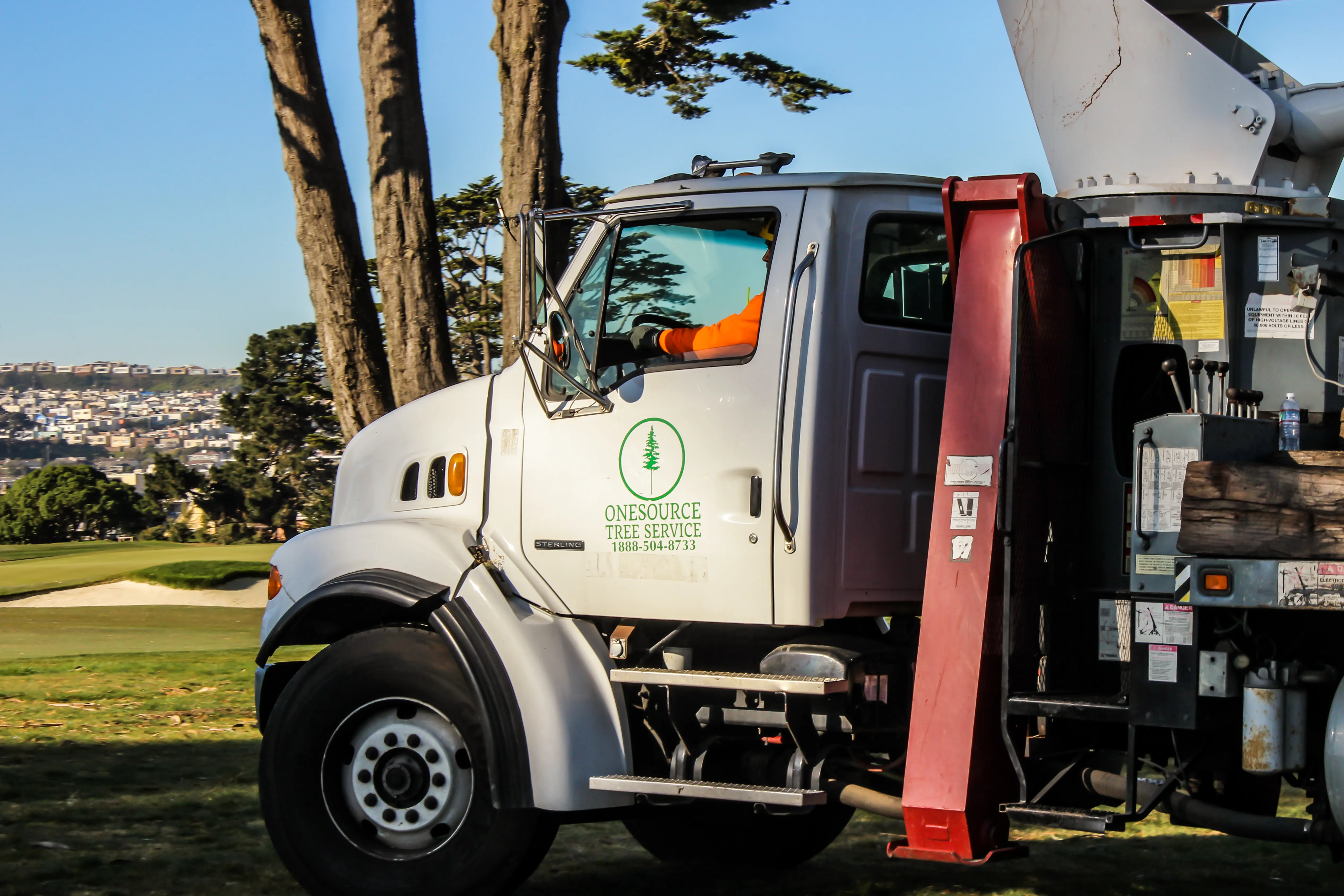 A Onesource Tree Service truck parked near a grassy area, with the company's logo visible on the door, and trees in the background.