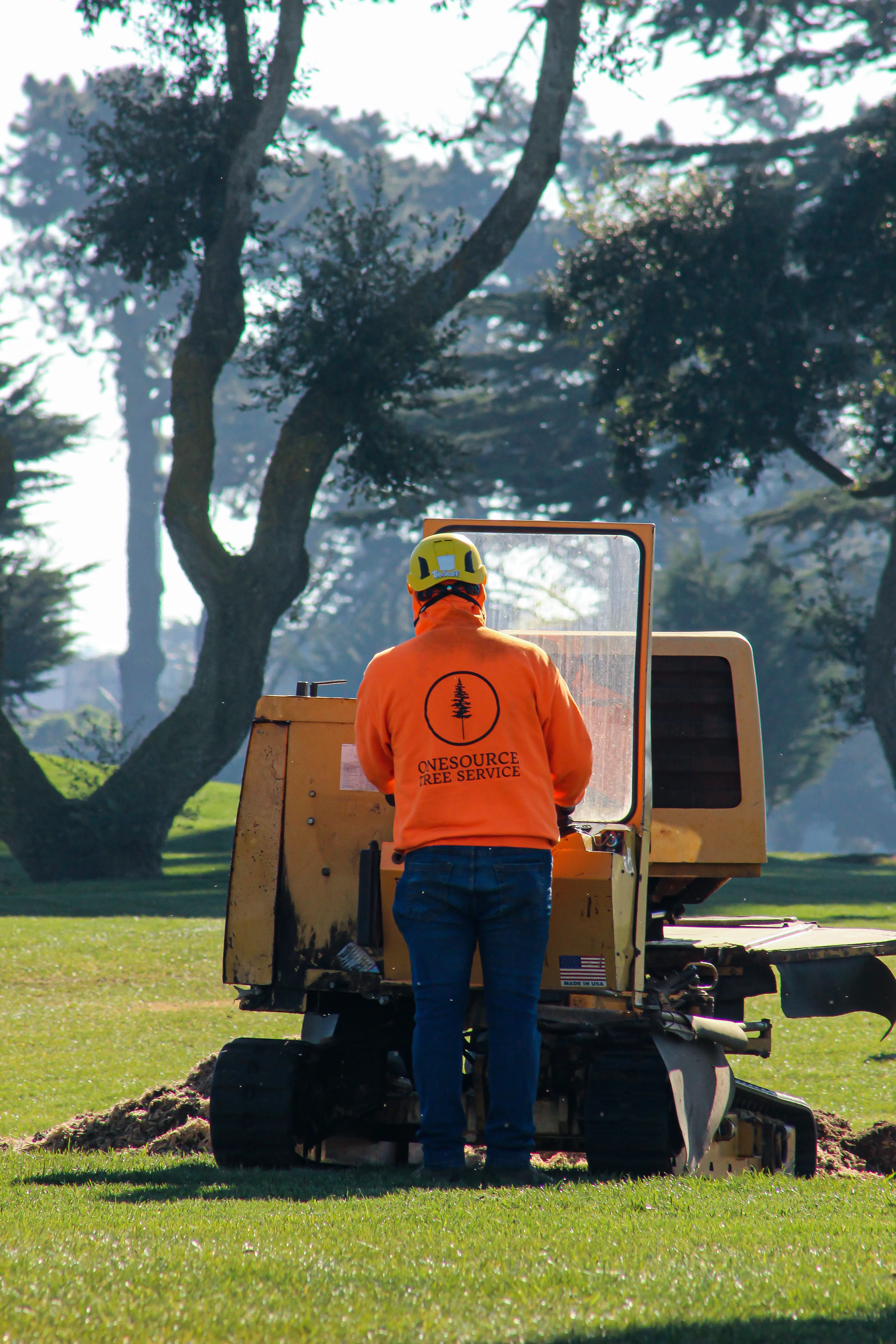 A member of Onesource Tree Service wearing an orange jacket operates a stump grinding machine on a grassy field in Newark.