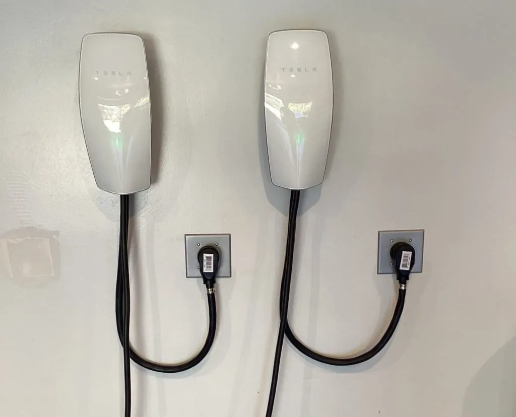 Two Tesla wall-mounted electric vehicle charging stations installed on a white wall, each with a connected charging cable.