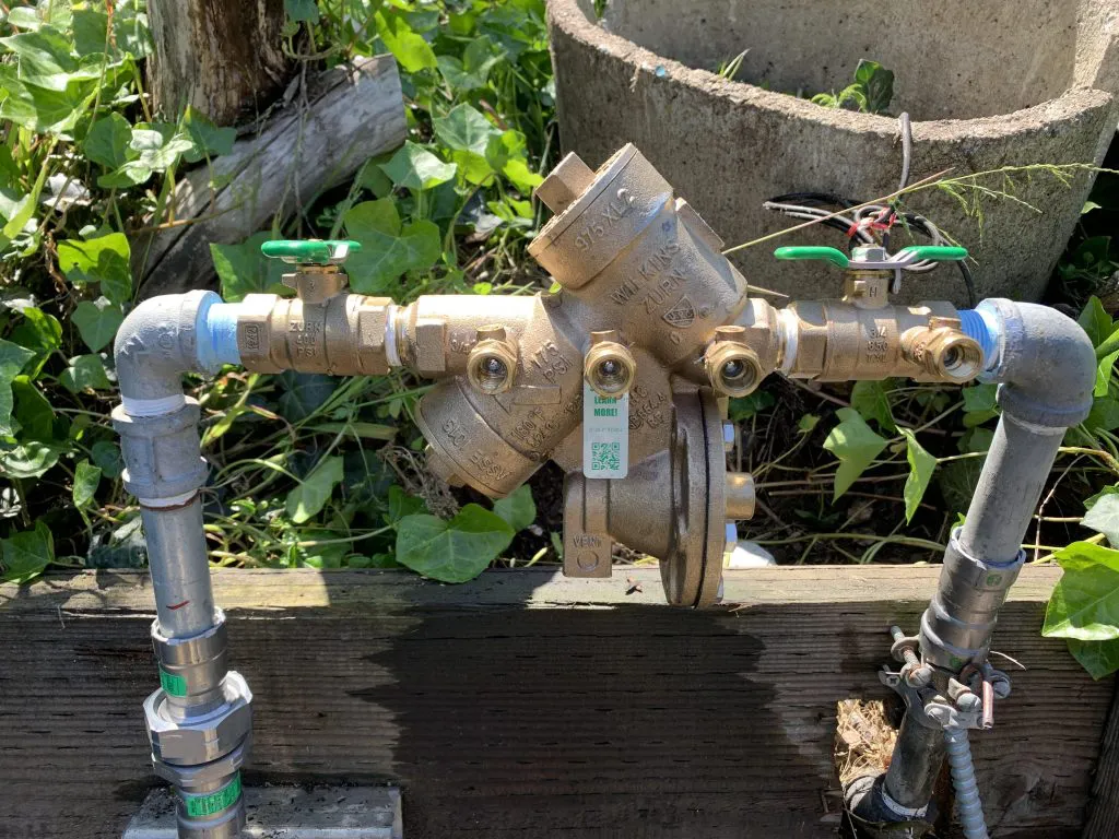 A brass backflow preventer with green-handled valves, part of a plumbing system, surrounded by greenery, installed by Ongaro & Sons in Santa Rosa.