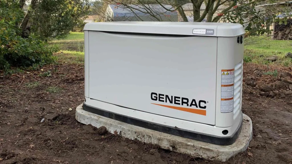 Generac generator installed on a concrete pad in a garden, surrounded by trees and grass.