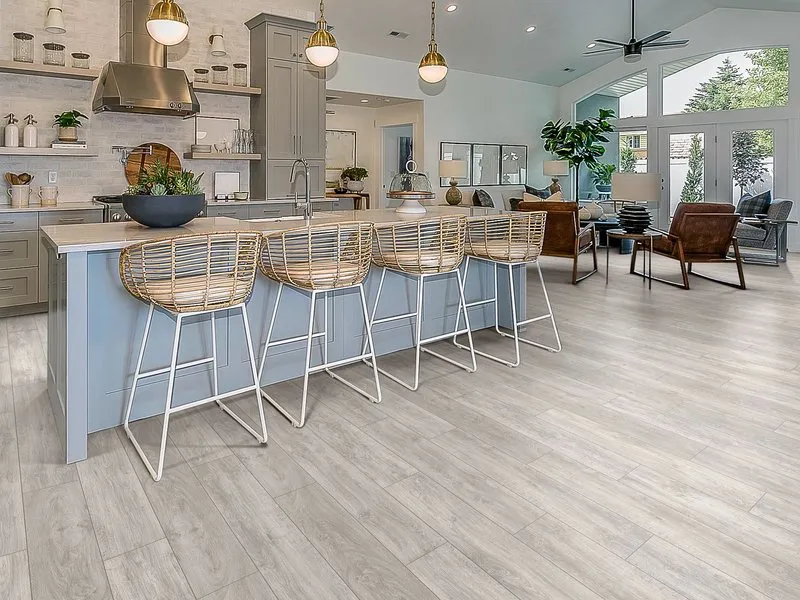 A modern kitchen with light wood flooring, featuring a center island with wicker bar stools, contemporary lighting, and an open living area in the background.