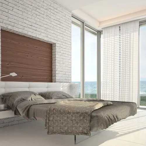 A modern bedroom with a bed featuring grey bedding, a white brick accent wall, large windows with sheer curtains, and an ocean view.