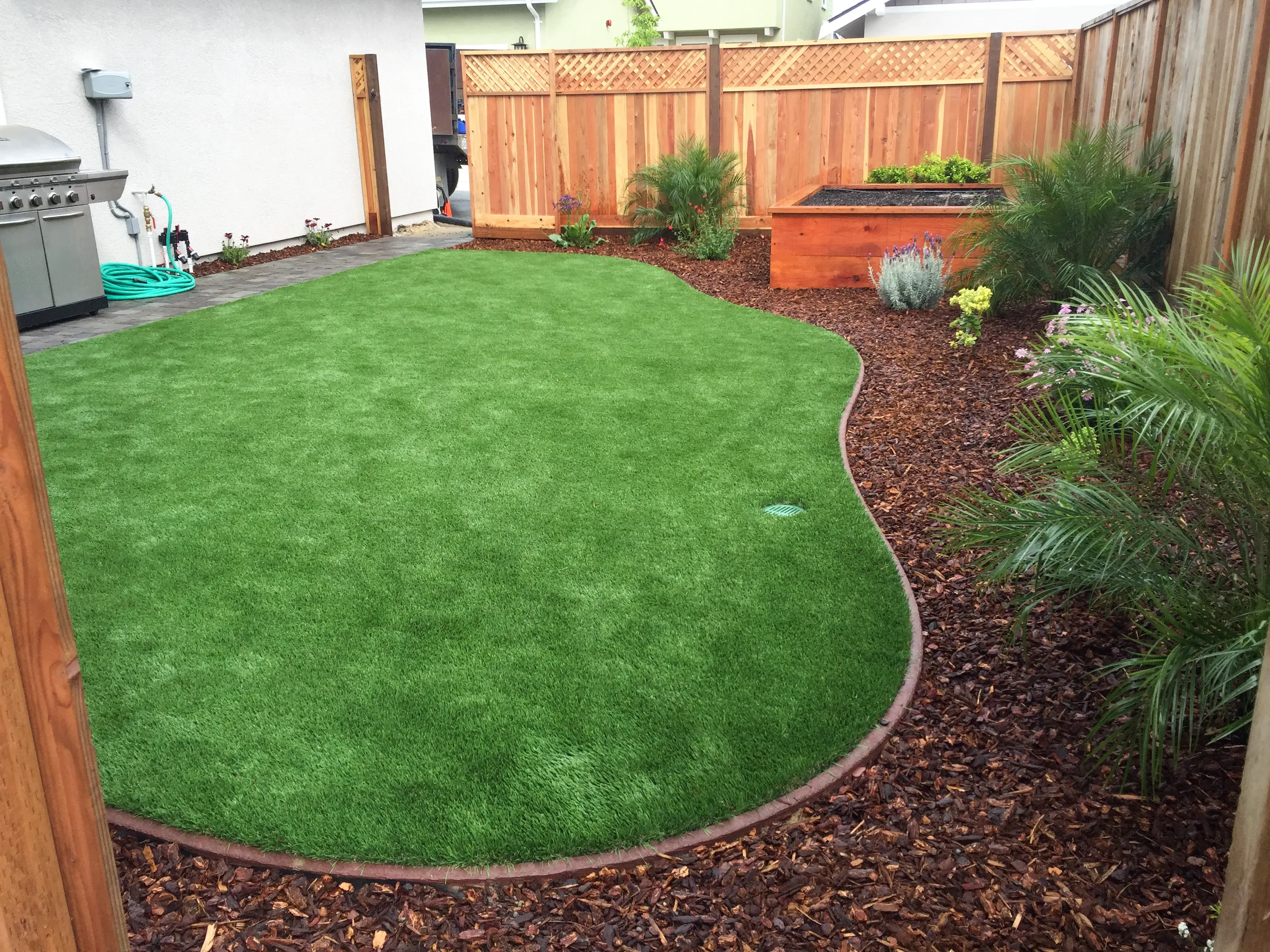 A well-maintained backyard featuring a lush green lawn, bordered by a mulch-covered garden with small plants and a wooden fence, alongside a barbecue grill, crafted by Pacific Landscaping & Tree Service Inc. in Pacific Grove.