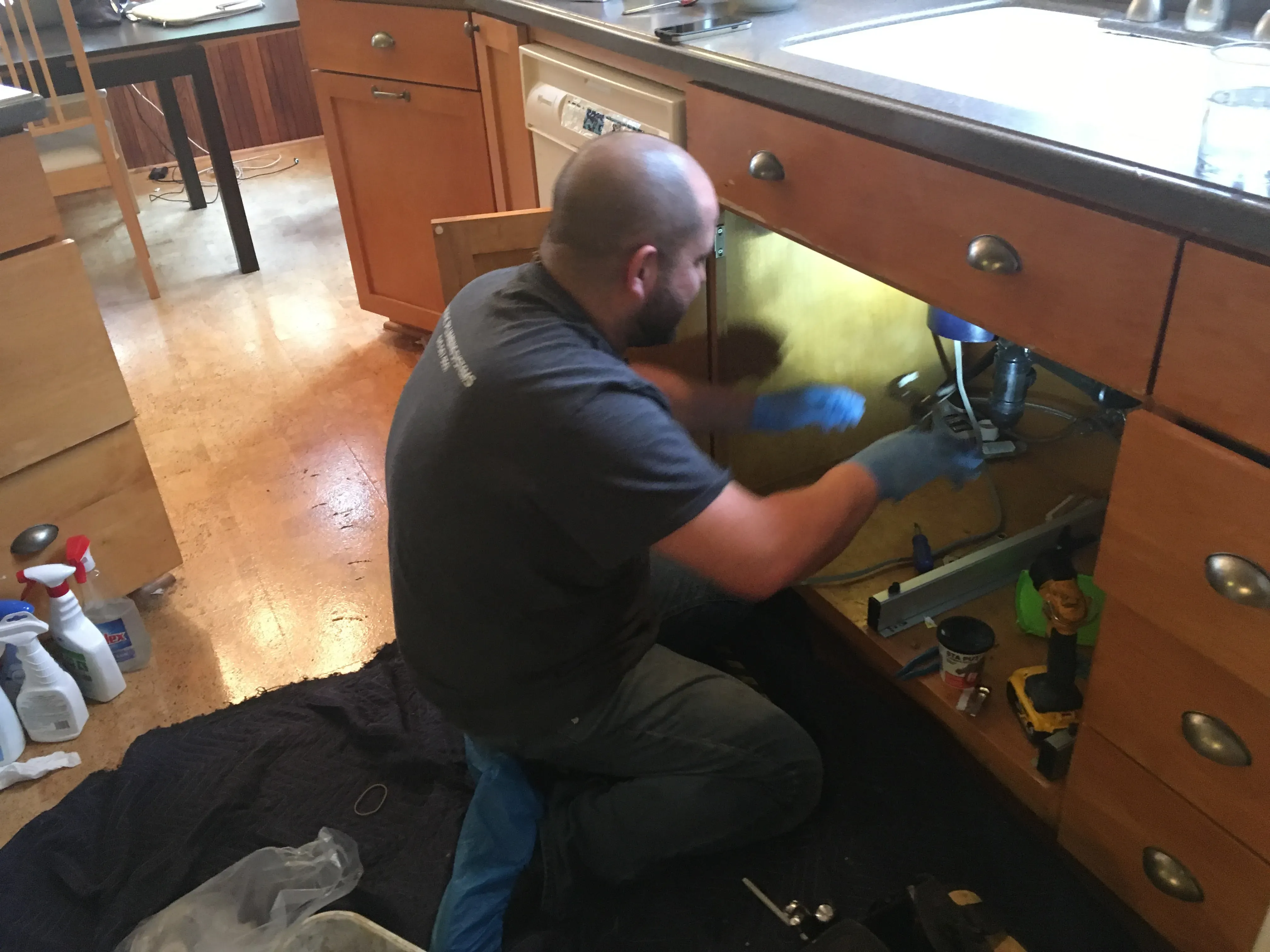 A plumber from Pacific Plumbing Systems in San Leandro is fixing pipes under a kitchen sink, using tools and wearing gloves for safety.