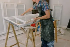 A person in a plaid shirt and apron painting a window frame on sawhorses in a workshop.