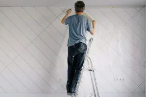 A person standing on a ladder meticulously painting a patterned wall.