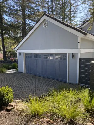 A modern, light gray garage door with windows at the top, surrounded by a landscaped driveway and greenery.