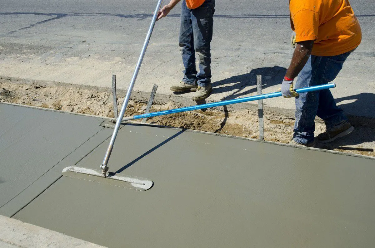 Two construction workers from Paradigm Concrete & Construction, Inc. in Antioch are smoothing freshly poured concrete on a sidewalk using long-handled tools.