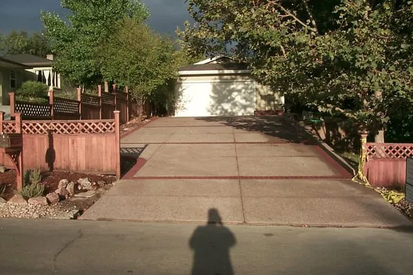 A well-constructed driveway in Antioch, featuring clean lines and robust design by Paradigm Concrete & Construction, Inc., bordered by wooden fences and lush trees.