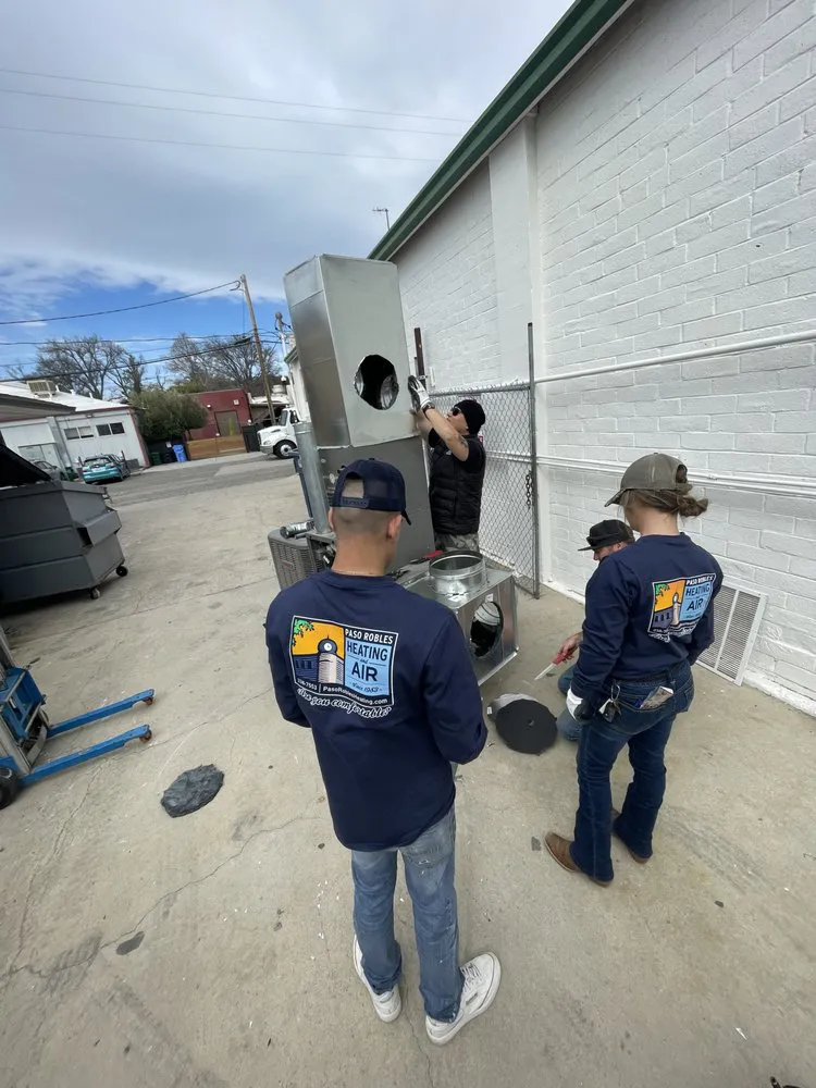 Technicians from Paso Robles Plumbing, Heating, and Air working on assembling a large metal unit outdoors, wearing company-branded clothing.