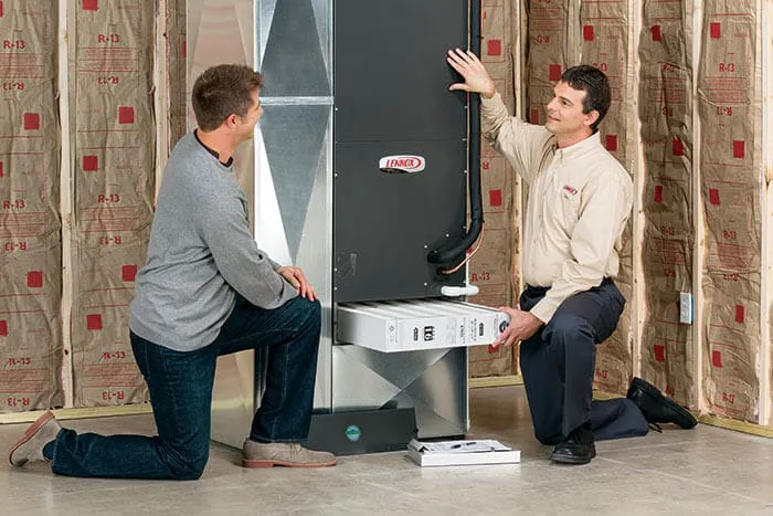 "A technician from Perfect Star Heating and Air Conditioning in Concord discusses furnace maintenance with a customer, both kneeling beside a furnace unit in an unfinished room."
