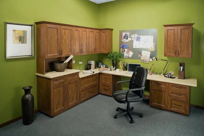 A home office featuring dark wooden cabinets and desks against a green wall, with a black chair and various office items organized on the surfaces, designed by PremierGarage in San Mateo.