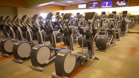 A row of modern elliptical machines lined up in a well-lit gym at Prime Time Athletic Club in Burlingame, with televisions mounted on the wall above.