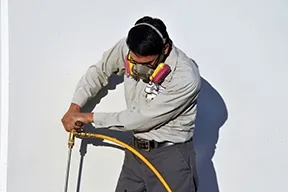 A technician from Proven Termite Solutions in San Jose using a hose and wearing protective gear to apply pest control treatment against a white wall.