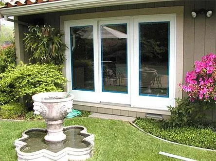 Three large white-framed glass doors opening to a lush garden with a fountain and vibrant plants, installed by Quality Windows & Doors, Inc. in Pleasanton.