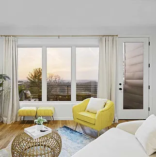 A modern living room featuring large windows and a glass door, showcasing an expansive outdoor view. Bright yellow furniture and minimalistic decor add a touch of elegance.