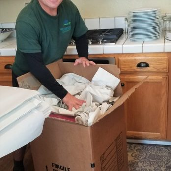 "A person from Quantum Moving carefully packs dishes into a large cardboard box, surrounded by packaging materials, in a kitchen in Martinez."