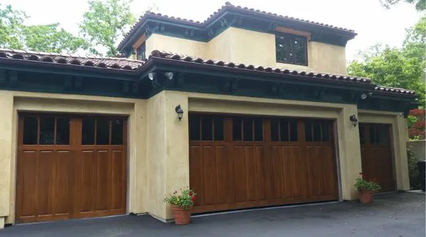 A house with three wooden garage doors and a beige exterior, featuring a dark roof and small potted plants.