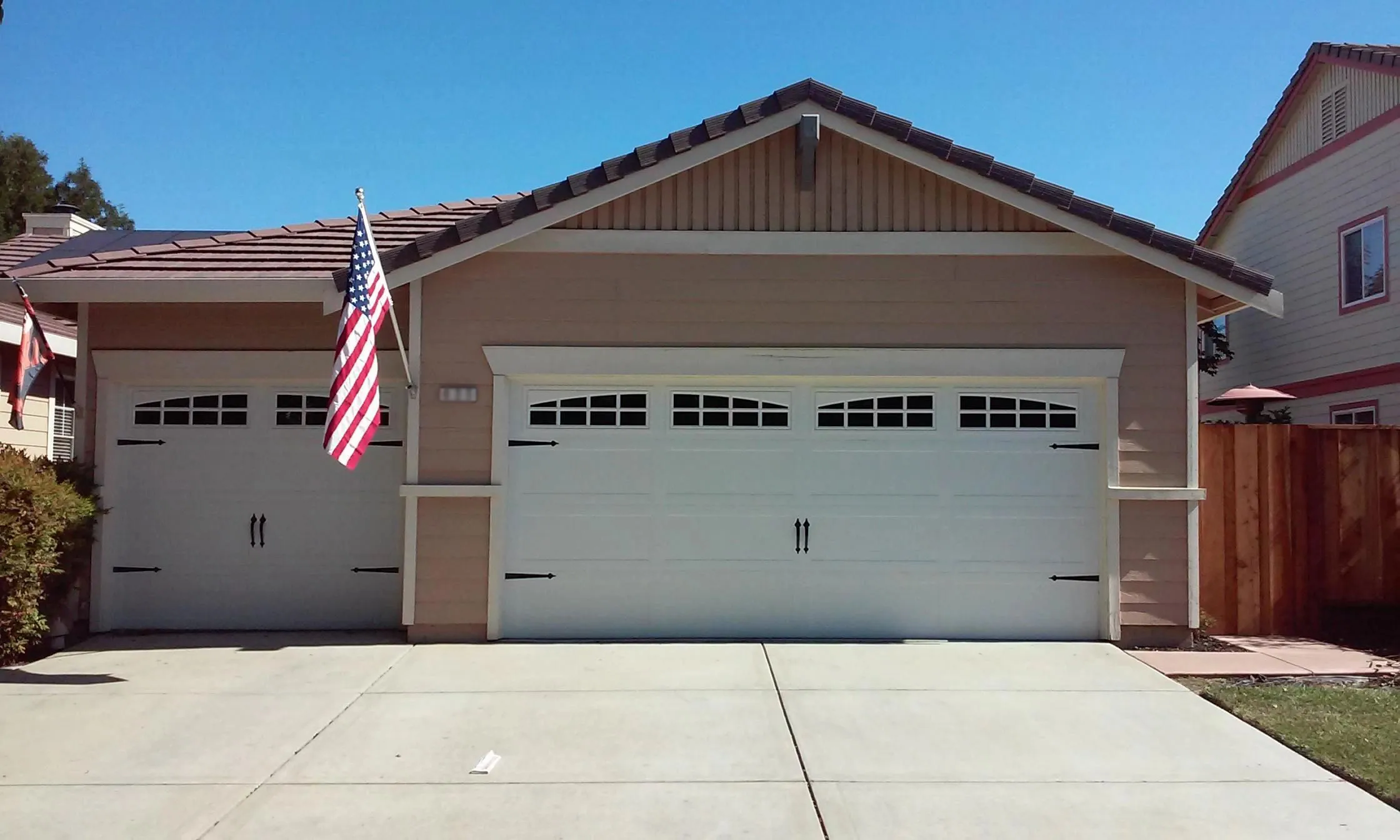 A tan house in Byron featuring a white double garage door with decorative windows and an American flag displayed on the side, installed by R & P Garage Doors.