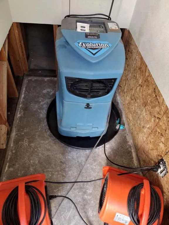 A dehumidification setup by Reactic Restoration in San Pablo, featuring a blue dehumidifier and two orange fans in a partially enclosed area with exposed wall sections.