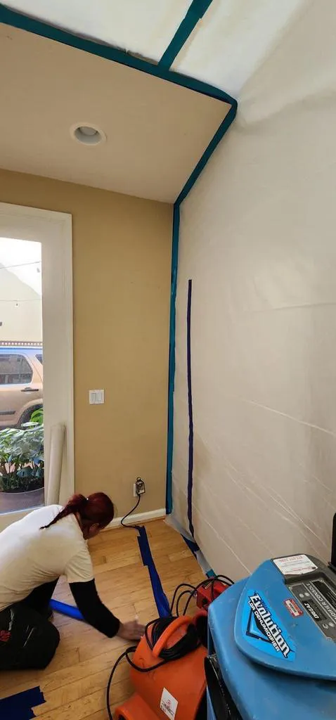 A person tapes plastic sheeting to a wall and floor corner, preparing the area for restoration work, with machinery from Reactic Restoration visible in the foreground.