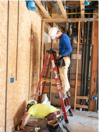 Two electricians from Reyff Electric, Inc. working inside a building under construction in Rohnert Park, one on a ladder and the other crouching, both wearing safety gear.