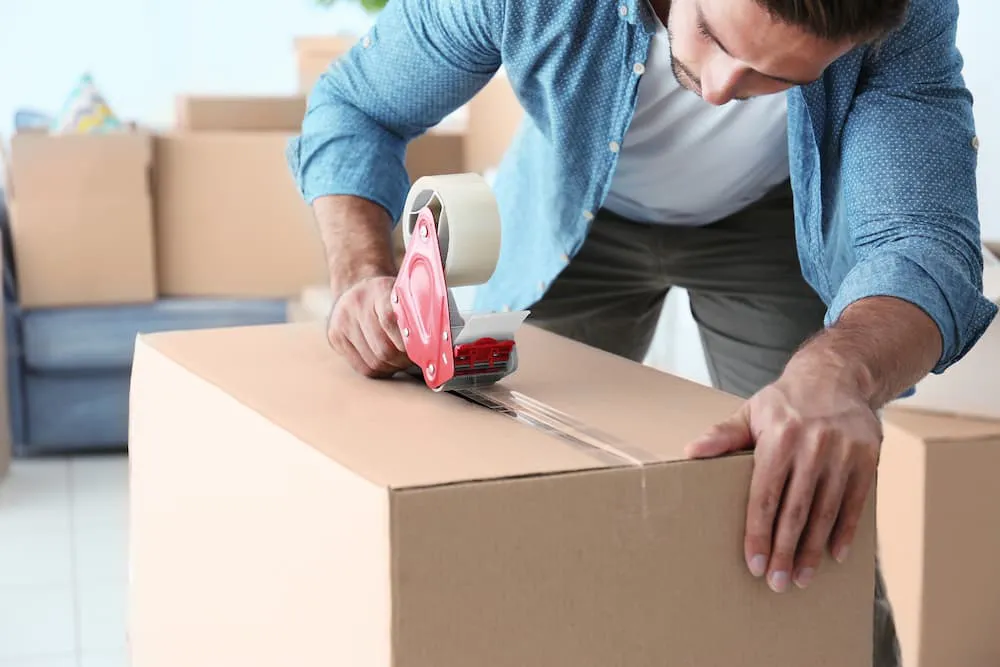 A person sealing a cardboard box with tape, surrounded by other packed boxes, preparing for a move.