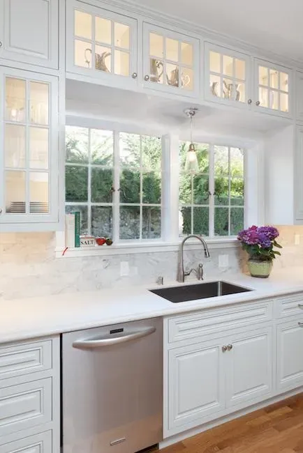 A brightly lit kitchen featuring white cabinets, a stainless steel dishwasher, and a window with greenery outside. There is a sink below the window and a potted plant on the counter.