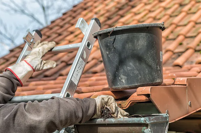 A worker wearing gloves cleans a gutter on a tiled roof with a bucket and ladder nearby.