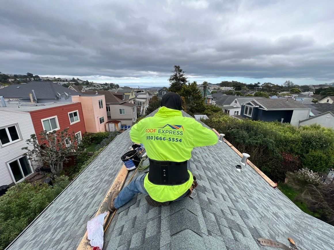A person wearing a neon green jacket, with "Roof Express" written on the back, works on a rooftop in a residential area with cloudy skies in San Francisco.