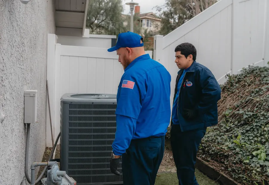 Two Rooter Hero Plumbing technicians in blue uniforms examining an outdoor HVAC unit next to a building in Martinez.