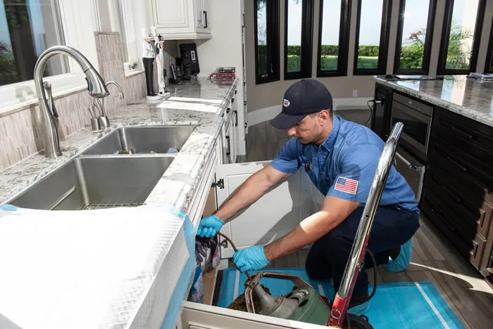 A plumber from Rooter Hero Plumbing working under a kitchen sink, wearing a blue uniform and gloves, demonstrating professional repair techniques in a well-lit kitchen.