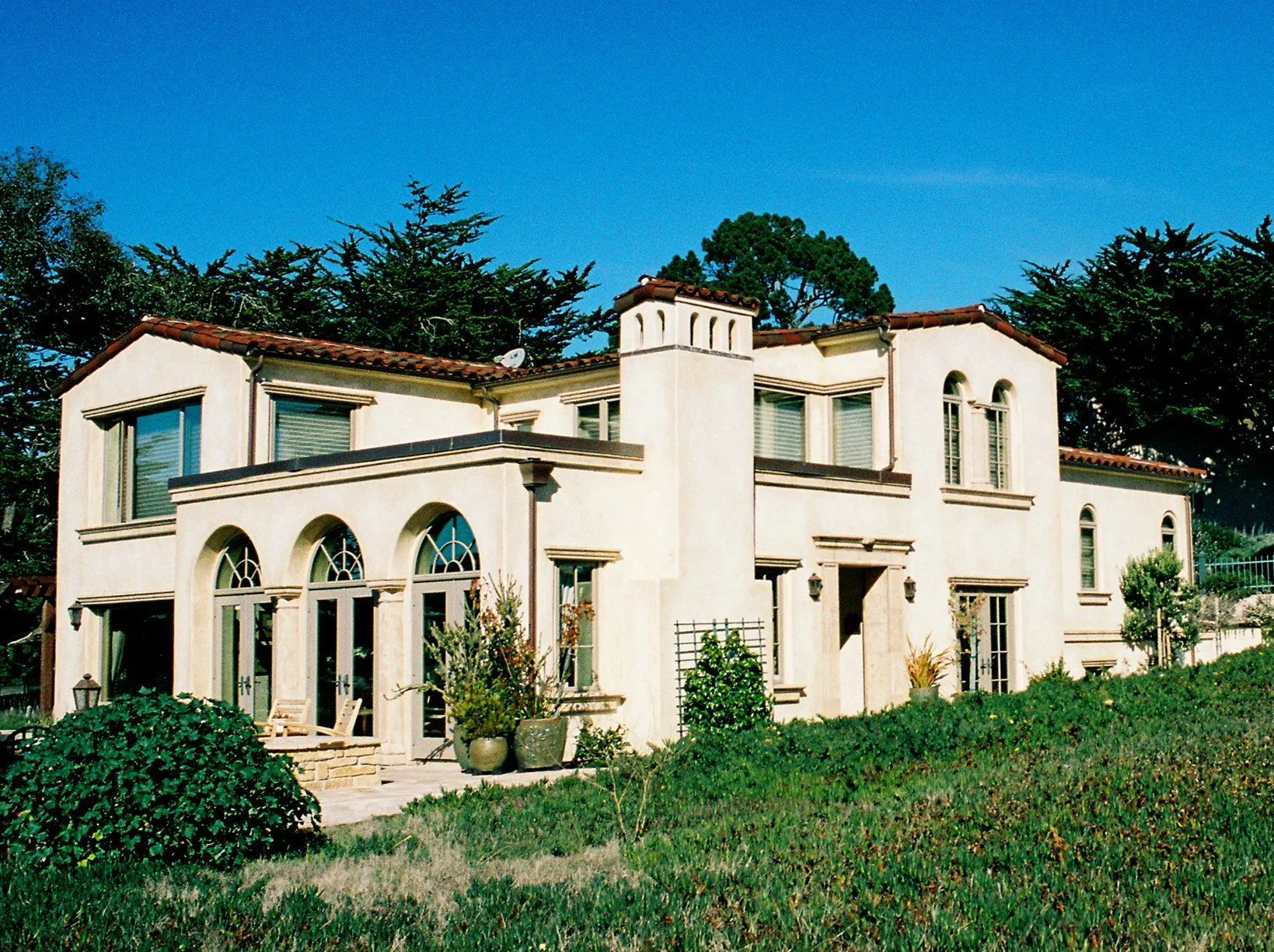A Mediterranean-style house with arched windows and terracotta roof, surrounded by greenery under a clear blue sky.