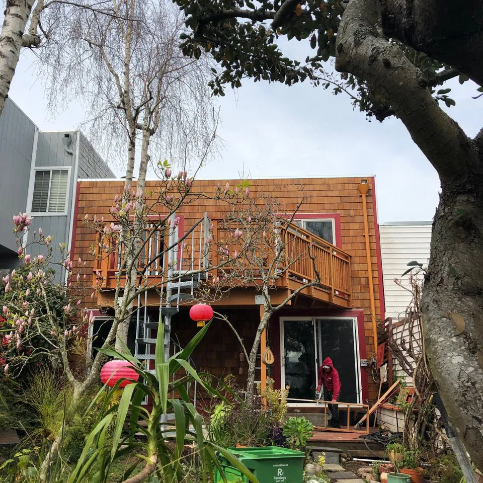 A wooden house exterior with a balcony, featuring trees and plants in the foreground, showcasing craftsmanship by SF Craftsman, Inc. in San Francisco.