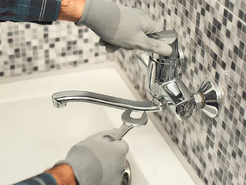 "A plumber from San Jose Plumbing, Inc. uses a wrench to repair a silver faucet against a tiled wall."