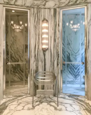 A luxurious bathroom setting featuring two marble shower doors with elegant lighting and a sleek metallic chair, showcasing Schicker Luxury Shower Doors, Inc. in Concord.