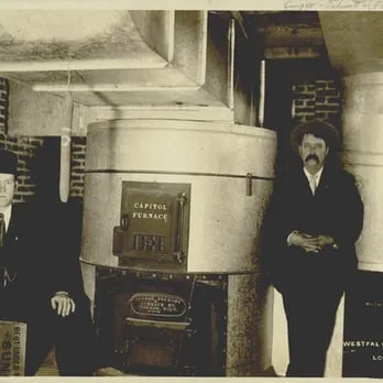 Sepia-toned photo of two men standing beside a large Capitol furnace in an industrial setting, representing early heating technology.
