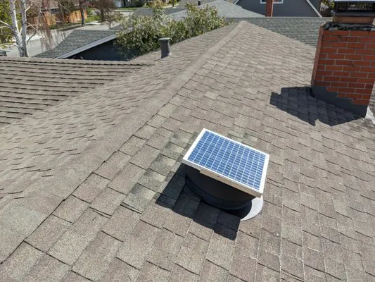 Roof with shingles and a small solar panel installed on a vent in a residential area.