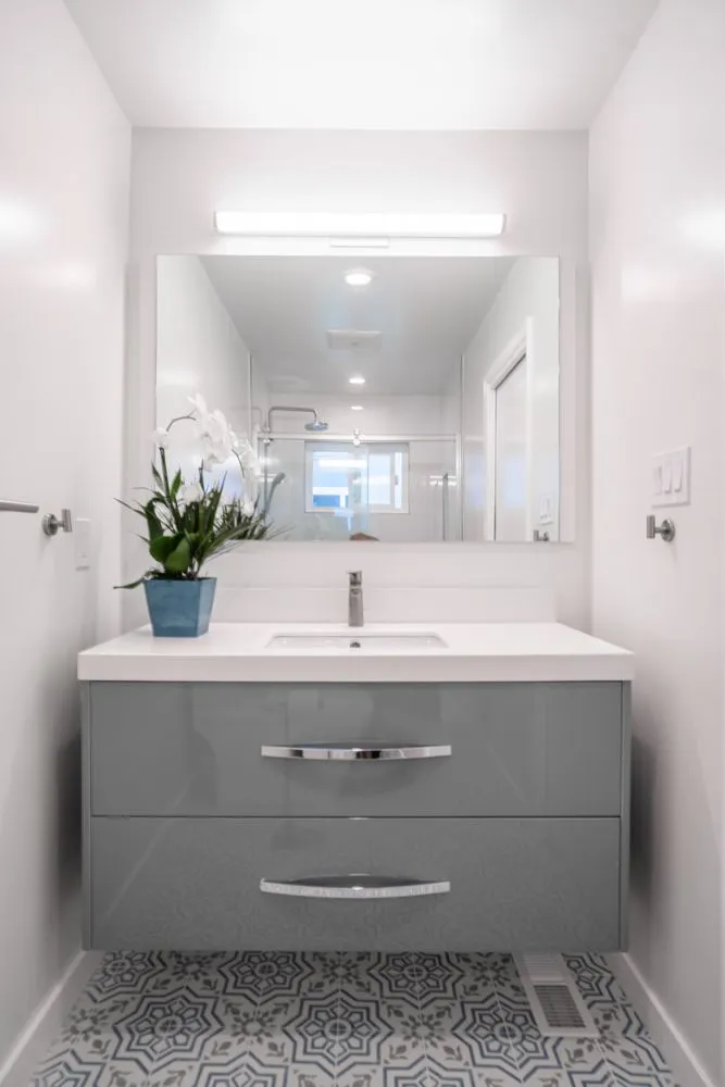 Modern bathroom interior featuring a sleek gray vanity with large drawers, a white countertop, a rectangular mirror, and decorative patterned floor tiles, designed by Simple Builders in Winnetka.