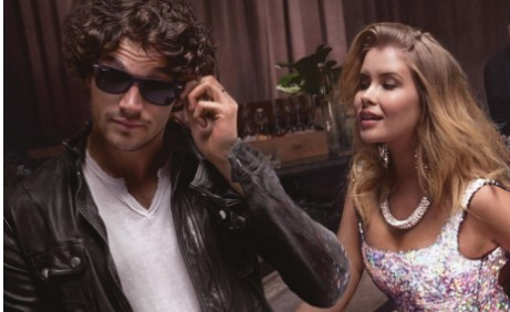 A man in sunglasses and a leather jacket adjusts his eyewear next to a woman in a sequined dress, both appearing stylish and confident.