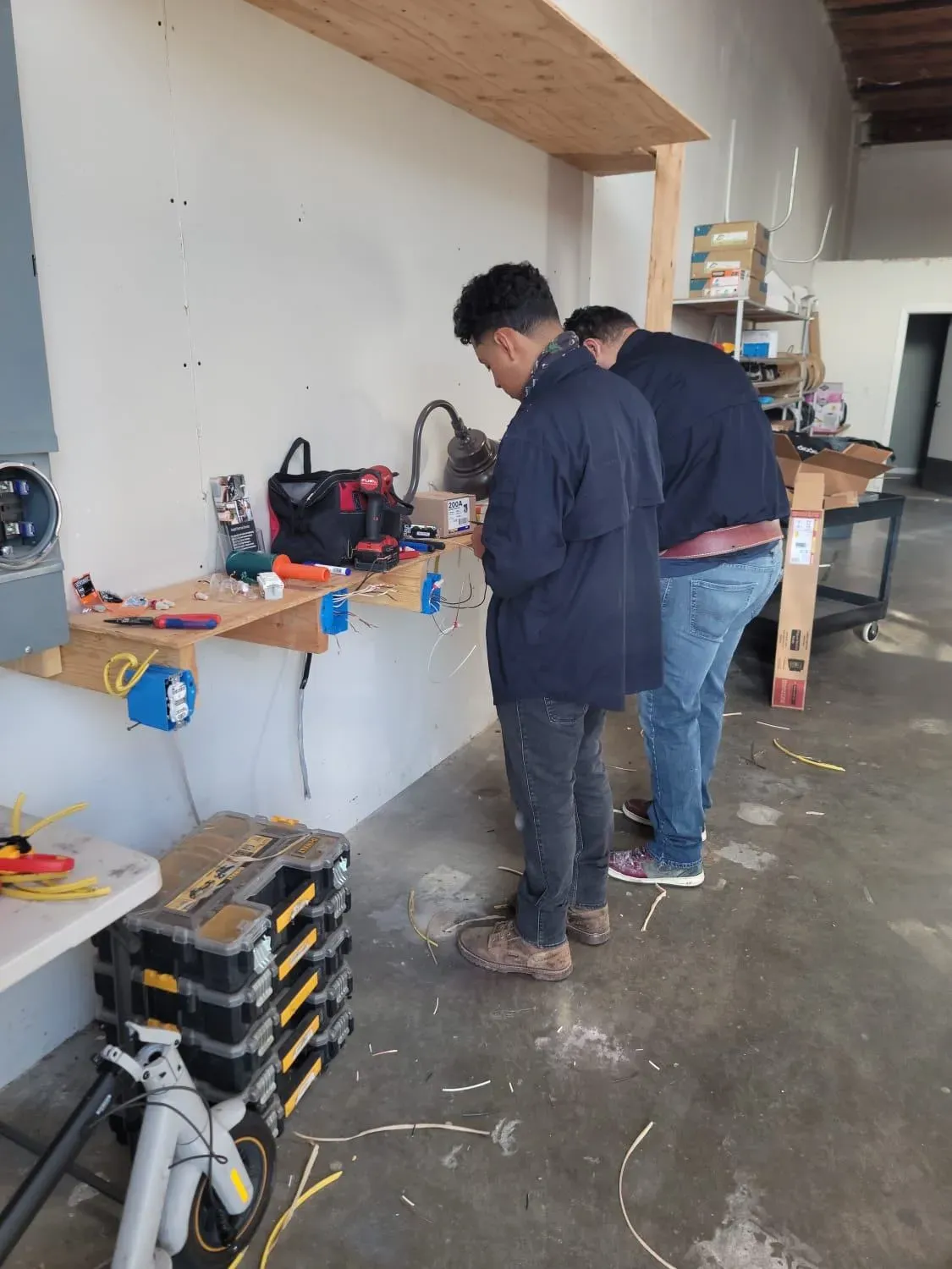 Two workers from Skylight Electrical Service in Fairfield are engaged in electrical tasks, surrounded by tools and wires in a workshop setting.