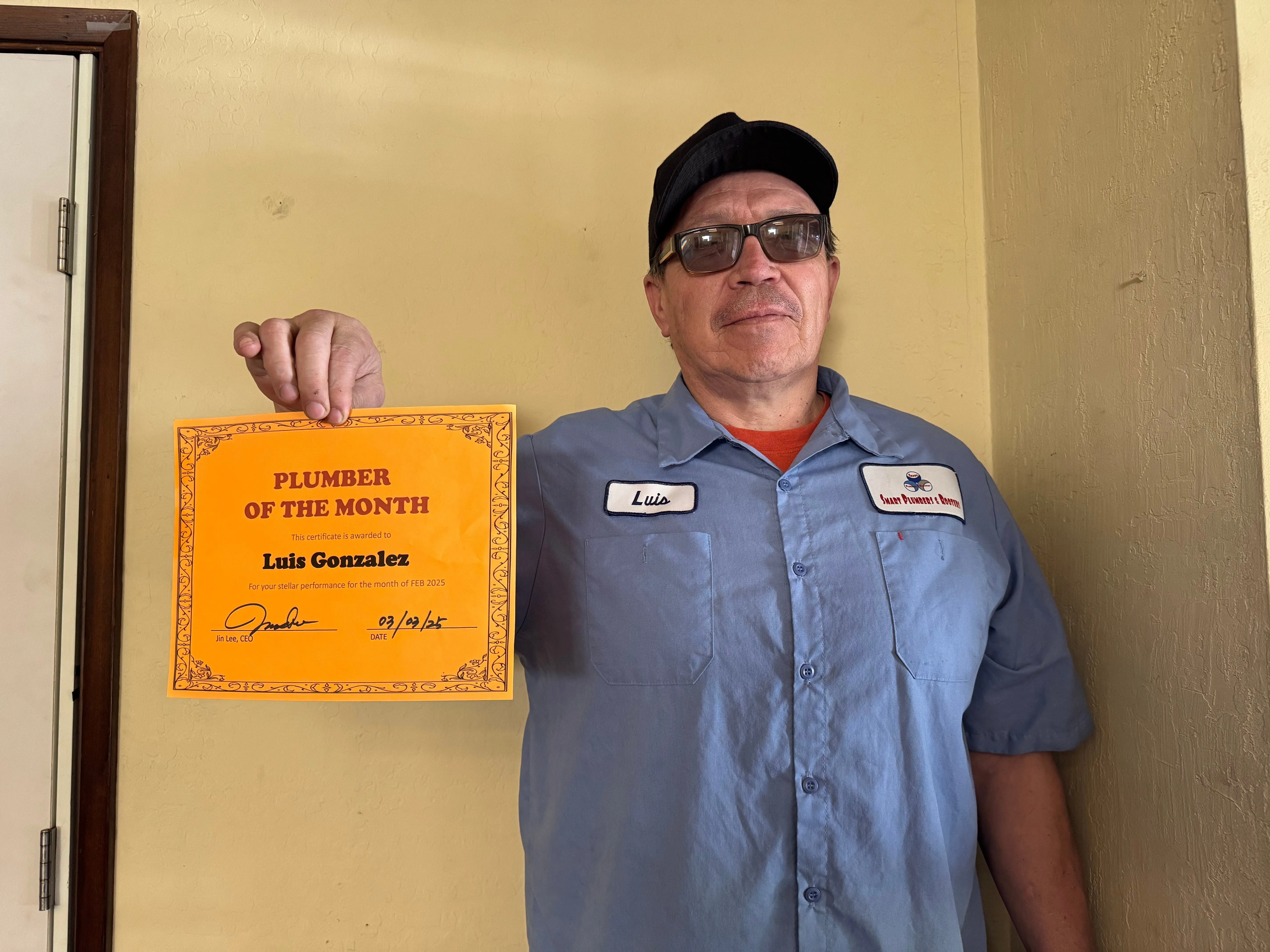 A man wearing a blue shirt with the Smart Plumbers, Inc. logo stands holding a "Plumber of the Month" certificate.