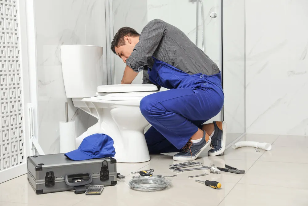 A plumber in blue overalls working on a toilet in a bathroom, surrounded by a set of tools, including a toolbox, at Smart Plumbers, Inc., Santa Clara.